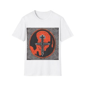 May include: White t-shirt featuring a circular graphic with a black cross design on an orange background. The graphic is set against a textured gray backdrop. The shirt is made of a soft, lightweight material.
