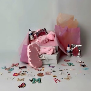 Puede incluir: Una caja de regalo blanca llena de artículos de cuidado personal rosas, incluyendo una máscara para dormir, una diadema y productos de belleza. Confeti decorativo y adornos con forma de letras están esparcidos alrededor de la caja.