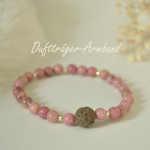 Könnte beinhalten: Ein rosa Perlenarmband mit einer strukturierten braunen Perle in der Mitte. Das Armband besteht aus kleinen, runden, rosa Perlen und silberfarbenen Abstandshaltern. Der Text "Duftträger-Armband" steht in Kursivschrift über dem Armband.