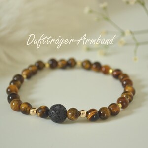 Könnte beinhalten: Ein Armband aus Tigeraugenperlen und goldfarbenen Perlen. Das Armband enthält eine dunkelgraue, poröse Lavasteinperle. Der Text "Duftträger-Armband" ist kursiv geschrieben.