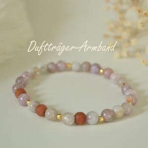 Könnte beinhalten: Ein Perlenarmband mit dem Text "Duftträger-Armband". Das Armband hat eine Mischung aus hellvioletten, weißen und braunen Perlen, die mit kleinen goldfarbenen Abstandshaltern durchsetzt sind. Die Perlen sind zu einer kreisförmigen Form aufgereiht.