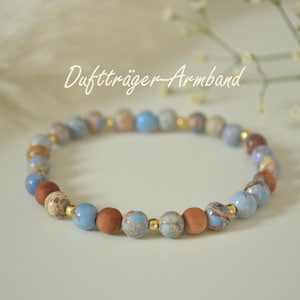 Könnte beinhalten: Ein Armband mit blauen, braunen und beigen Kugeln. Es enthält kleine goldfarbene Abstandshalter und den Text "Duftträger-Armband" in einer Schreibschrift.