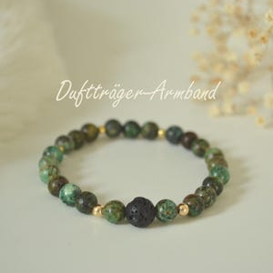 Könnte beinhalten: Ein Armband mit grünen und braunen Steinperlen, unterbrochen von goldfarbenen Perlen. Eine einzelne schwarze, poröse Steinperle ist zentriert. Das Armband ist mit "Duftträger-Armband" in Schreibschrift beschriftet.