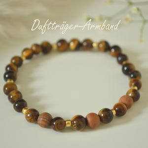 Könnte beinhalten: Ein Armband mit Tigerauge-, Holz- und rotbraunen Perlen. Goldfarbene Abstandshalter sind eingearbeitet. Der Text "Duftträger-Armband" ist in einer Schreibschrift dargestellt.