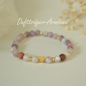 Könnte beinhalten: Armband mit goldenem Herzanhänger. Es besteht aus hellvioletten, rosa und braunen Perlen, die durch kleine goldfarbene Perlen unterbrochen werden. Der Text "Duftträger-Armband" ist kursiv geschrieben.