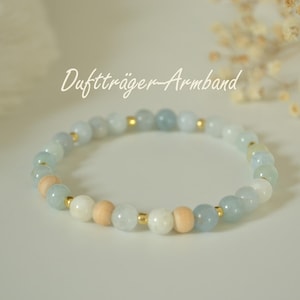 Könnte beinhalten: Ein Armband mit hellblauen, weißen und Holzperlen, unterbrochen von kleinen goldfarbenen Akzenten. Das Armband ist auf einem Gummiband aufgefädelt. Der Text "Duftträger-Armband" ist in Schreibschrift geschrieben.