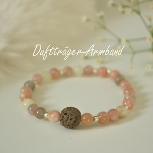 Könnte beinhalten: Ein Armband mit rosa, weißen und grauen Perlen und einer großen braunen Lavasteinperle. Das Armband hat goldfarbene Abstandshalter und den Text "Duftträger Armband" in Schreibschrift.