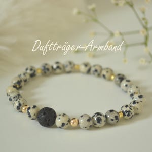 Könnte beinhalten: Ein Perlenarmband mit Dalmatiner-Jaspis-Perlen, mit schwarzen Flecken und goldfarbenen Akzenten. Das Armband enthält eine schwarze Lavasteinperle und ist mit "Duftträger-Armband" beschriftet.