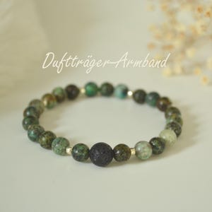Könnte beinhalten: Ein Perlenarmband mit grünen und schwarzen Steinen, darunter eine zentrale schwarze Lavaperle. Auf dem Armband steht "Duftträger-Armband". Die Perlen sind auf einer flexiblen Schnur aufgefädelt.