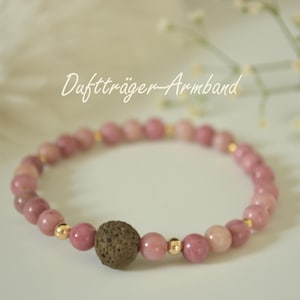 Könnte beinhalten: Ein rosa Perlenarmband mit einer braunen Lavasteinperle und goldfarbenen Akzenten. Das Armband besteht aus kleinen, runden, rosa Perlen. Der Text "Duftträger-Armband" ist in Kursivschrift oben im Bild geschrieben.