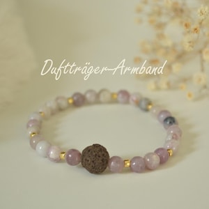 Könnte beinhalten: Ein Perlenarmband mit dem Text "Duftträger-Armband". Das Armband hat hellviolette und weiße Perlen, die mit kleinen goldfarbenen Abstandshaltern durchsetzt sind. Eine größere, strukturierte braune Perle befindet sich in der Mitte des Armbands.