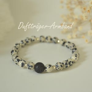 Könnte beinhalten: Armband mit Dalmatiner-Jaspis-Perlen mit schwarzen Punkten und einer zentralen schwarzen Lavasteinperle. Das Armband enthält den Text "Duftträger-Armband". Die Perlen sind auf einem elastischen Band aufgefädelt.