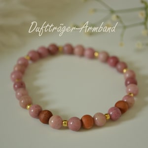 Könnte beinhalten: Ein rosa Perlenarmband mit goldenen Akzenten und Holzperlen. Das Armband hat eine Mischung aus rosa und braunen Perlen, die durch kleine goldfarbene Abstandshalter verbunden sind. Der Text "Duftträger Armband" ist sichtbar.