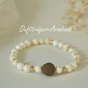 Könnte beinhalten: Weißes Perlenarmband mit goldenen Akzenten und einer dunkelbraunen, strukturierten Perle. Das Armband trägt den Schriftzug "Duftträger-Armband" in Schreibschrift. Das Armband besteht wahrscheinlich aus Natursteinperlen.