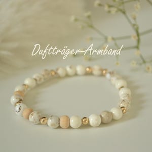 Könnte beinhalten: Ein Armband mit weißen, beigen und Holzperlen, unterbrochen von kleinen goldfarbenen Perlen. Das Armband wird auf einer weißen Oberfläche präsentiert, mit dem Text "Duftträger-Armband" im Bild.