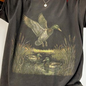 K&ouml;nnte beinhalten: Dunkelgraues T-Shirt mit einer detaillierten Illustration von Enten in einem Teich. Eine Ente ist im Flug, die Fl&uuml;gel ausgebreitet, &uuml;ber dem Wasser. Das Design umfasst Schilf und eine naturalistische Farbpalette, die eine Vintage-&Auml;sthetik erzeugt.