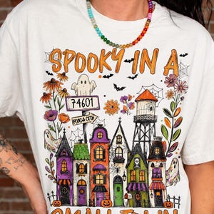 May include: An off-white t-shirt featuring a colourful Halloween-themed graphic of a small town with houses, a water tower, ghosts, flowers, and spiderwebs. The text reads "SPOOKY IN A SMALL TOWN" with a street sign labelled "74601 PONCA CITY".