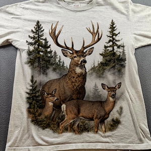 Peut inclure: Un t-shirt gris clair avec un motif d&eacute;taill&eacute; d'un cerf avec de grands bois, accompagn&eacute; de deux petits cerfs, sur fond de for&ecirc;t brumeuse avec des conif&egrave;res. Le t-shirt est fait d'une mati&egrave;re douce.