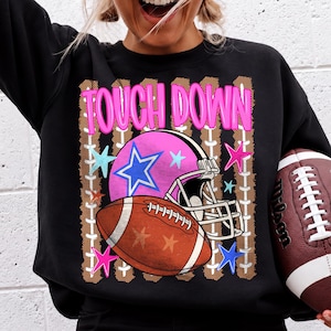 Peut inclure: Un sweat-shirt noir à col rond avec une impression graphique portant le texte TOUCH DOWN en lettres roses. Le motif comprend un casque de football américain rose avec une étoile bleue, un ballon de football marron et des étoiles colorées.