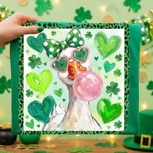May include: Green St. Patrick's Day themed t-shirt featuring a whimsical goose wearing heart-shaped sunglasses and a polka dot bow, blowing a bubblegum bubble. The design includes green hearts and shamrocks, with a leopard print border.