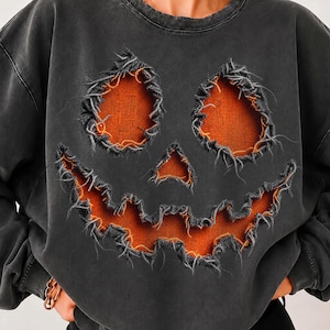 Op de afbeelding: Een antracietgrijze sweater met ronde hals en een grote, verweerde print van een pompoengezicht. Het ontwerp bestaat uit twee driehoekige ogen, een driehoekige neus en een grillige mond met gerafelde randen die een oranje stof eronder onthullen.