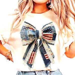 Op de afbeelding: Een crèmekleurig t-shirt met een grote, patriottische strik. De strik is gemaakt van denim, stof met patronen en kant, met elementen van de Amerikaanse vlag en het Vrijheidsbeeld. De strik is het middelpunt van het shirt.