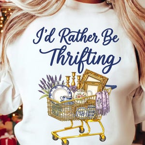 May include: White sweatshirt with the text "I'd Rather Be Thrifting" in blue script. A gold shopping cart graphic is filled with vintage items like plates, teacups, and picture frames. The cart also contains lavender and a purple scarf.