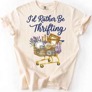 May include: Cream-colored t-shirt with the text "I'd Rather Be Thrifting" in navy blue script. A shopping cart graphic is filled with vintage items like plates, teacups, and a framed picture. The cart is yellow.
