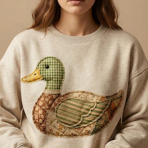 May include: A light beige sweatshirt featuring a large, appliqu&eacute;d duck design. The duck has a green and white checkered head, a yellow beak, and a body composed of various patterned fabrics in brown, green, and yellow hues. The design is quilted.