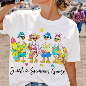 Op de afbeelding: Wit T-shirt met een afbeelding van cartoon ganzen in zomerkleding, inclusief zonnebrillen, hoeden en strandaccessoires. De tekst "Just a Summer Goose" staat eronder.