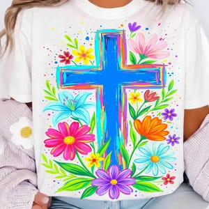 May include: White t-shirt featuring a large, vibrant blue cross with colorful brushstroke accents. The cross is surrounded by an array of bright flowers in pink, orange, yellow, and purple, with green leaves. The design has a watercolor effect.