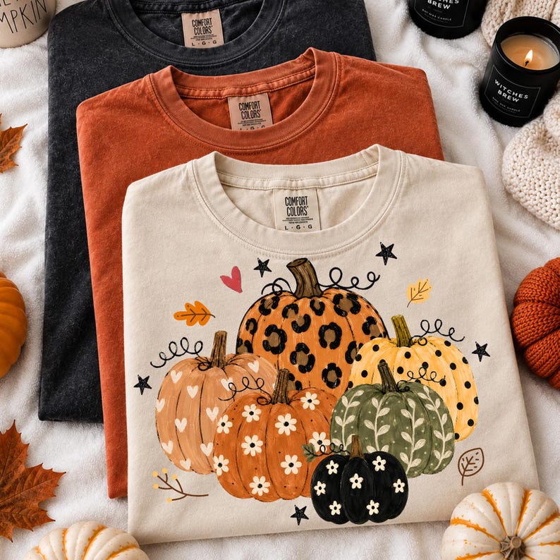 May include: Three crew neck t-shirts in black, burnt orange, and cream. The cream shirt features a graphic of five pumpkins with leopard print, polka dot, and floral patterns, accented by small hearts, stars, and leaves. Each shirt has a Comfort Colors label.