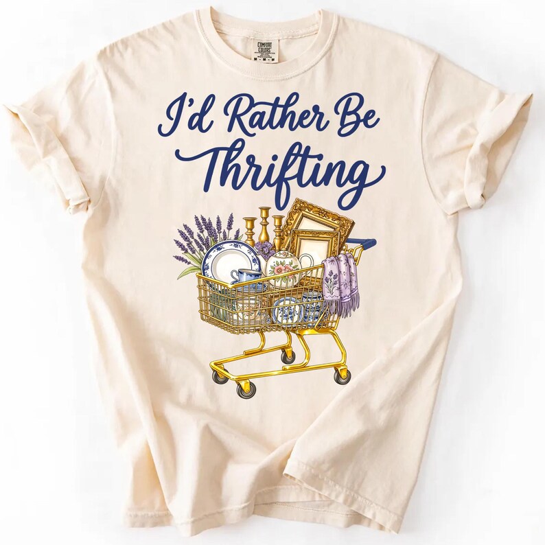 May include: Cream-colored t-shirt with the text "I'd Rather Be Thrifting" in navy blue script. A shopping cart graphic is filled with vintage items like plates, teacups, and a framed picture. The cart is yellow.