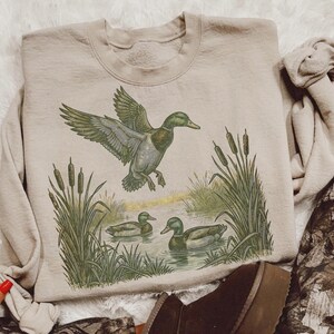 K&ouml;nnte beinhalten: Beiges Sweatshirt mit detaillierter Illustration von Enten in einer Feuchtgebietszene. Das Design zeigt eine Ente im Flug, Schilf und Enten, die im Wasser schwimmen. Das Sweatshirt hat eine neutrale Farbe und ist f&uuml;r den Freizeitgebrauch geeignet.
