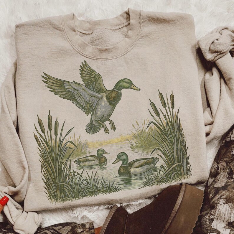 K&ouml;nnte beinhalten: Beiges Sweatshirt mit detaillierter Illustration von Enten in einer Feuchtgebietszene. Das Design zeigt eine Ente im Flug, Schilf und Enten, die im Wasser schwimmen. Das Sweatshirt hat eine neutrale Farbe und ist f&uuml;r den Freizeitgebrauch geeignet.
