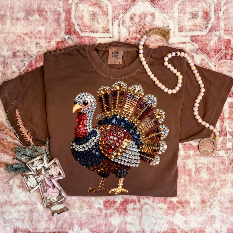 May include: A brown short-sleeve t-shirt featuring a graphic of a turkey composed of multicolored rhinestones. The turkey has a red wattle, a yellow beak, and a fanned tail made of gold, brown, and clear crystals. The shirt has a crew neckline.