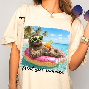 May include: Cream-colored t-shirt featuring a graphic of a sloth wearing heart-shaped sunglasses and floating on a pink donut in the ocean. The sloth is holding a cocktail. The text "feral girl summer" is below the graphic.