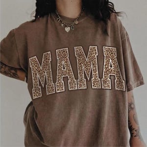 May include: A brown t-shirt with the word "MAMA" in large, block letters across the front. The letters are filled with a leopard print pattern and outlined in white. The shirt has short sleeves and a relaxed fit.