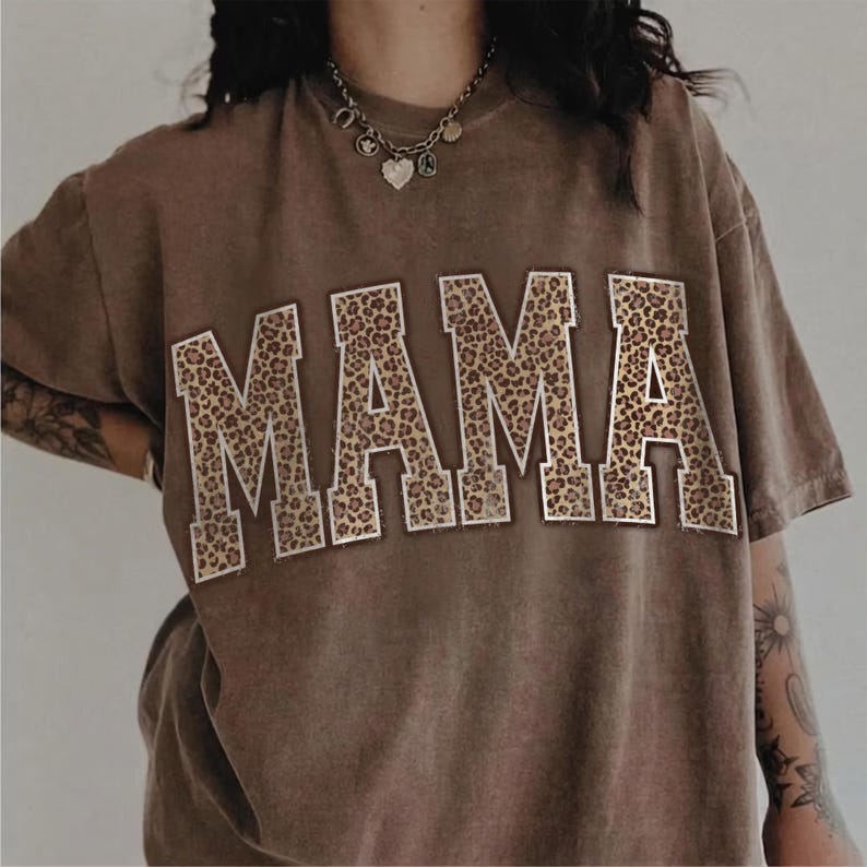 May include: A brown t-shirt with the word "MAMA" in large, block letters across the front. The letters are filled with a leopard print pattern and outlined in white. The shirt has short sleeves and a relaxed fit.