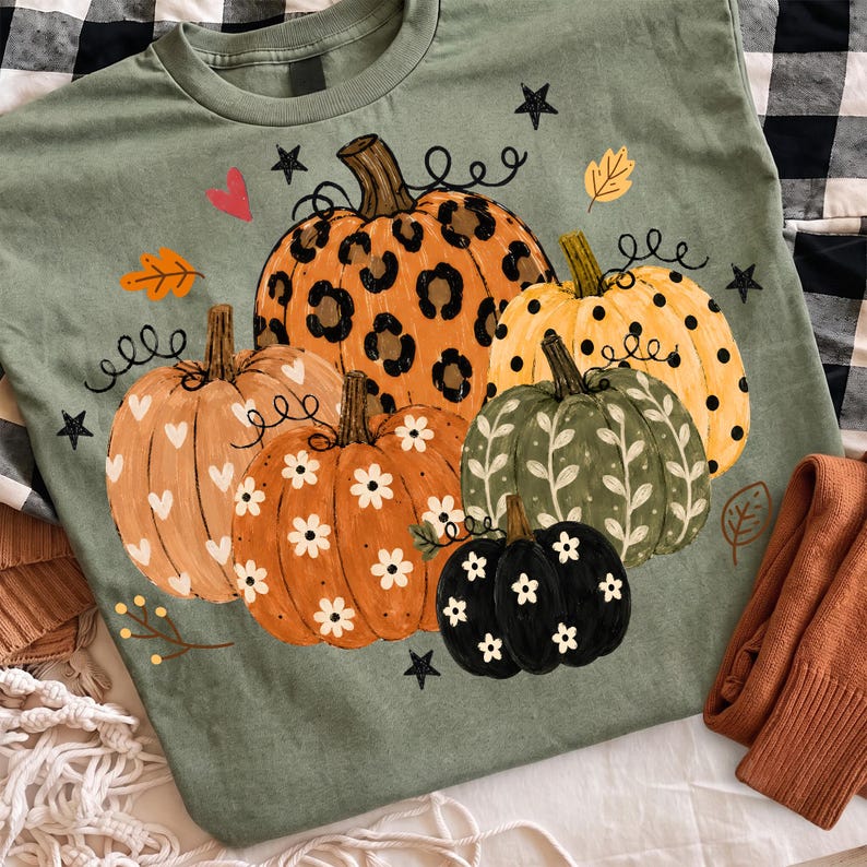 May include: A sage green t-shirt featuring a graphic print of five pumpkins in various colors and patterns, including leopard print, polka dots, and floral designs, accented with small black stars, hearts, and autumn leaves scattered around.