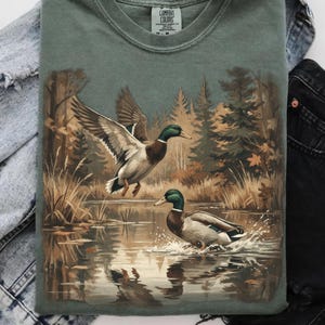 Vintage mallard ducks artwork on green sweatshirt, showing two ducks in a forest wetland scene with water reflections, autumn trees, and retro 90s outdoor hunting style digital design.