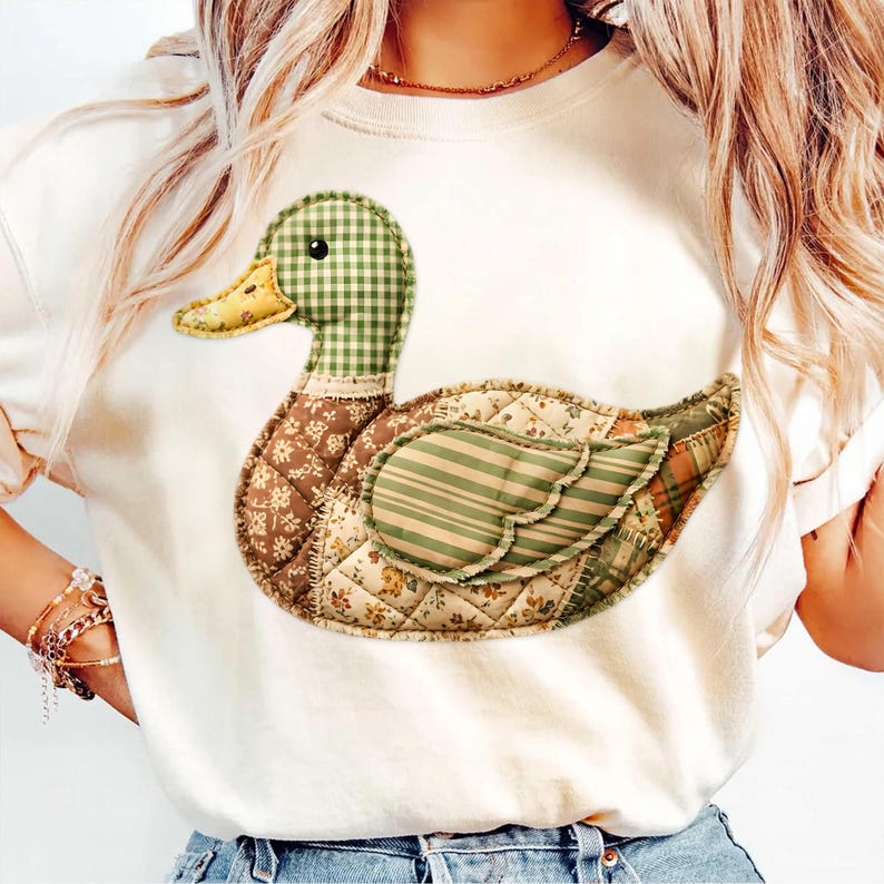 May include: Cream-colored t-shirt featuring a patchwork duck design. The duck has a green and white checkered head, a yellow beak, and a body composed of various patterned fabrics in shades of brown, green, and cream.