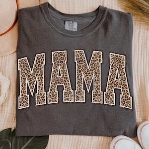 May include: Dark gray t-shirt with the word "MAMA" in large, bold letters across the front. The letters are filled with a leopard print pattern and outlined in a light brown color. The shirt is a Comfort Colors brand.