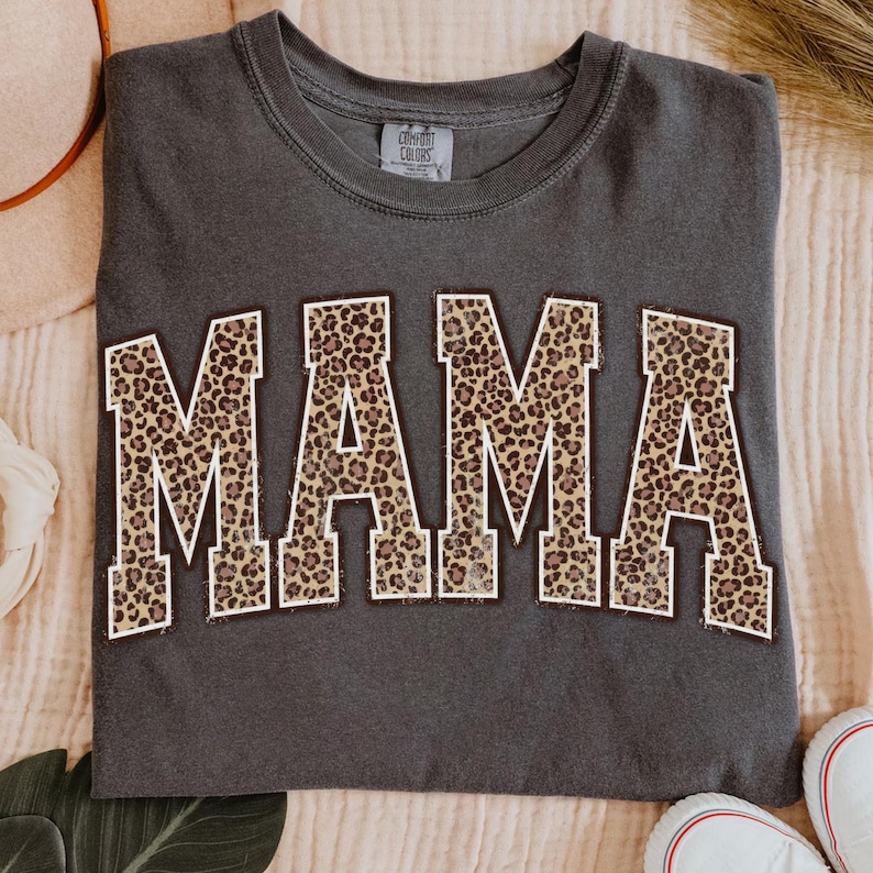 May include: Dark gray t-shirt with the word "MAMA" in large, bold letters across the front. The letters are filled with a leopard print pattern and outlined in a light brown color. The shirt is a Comfort Colors brand.