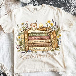 Puede incluir: Camiseta color crema con una pila de libros con t&iacute;tulos como "Wildflower Chronicles" y "Botanical Wonders". Una taza de t&eacute;, una vela y gafas est&aacute;n encima de los libros, rodeados de ilustraciones florales. El texto "Just One More Chapter" est&aacute; debajo.