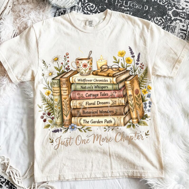Puede incluir: Camiseta color crema con una pila de libros con t&iacute;tulos como "Wildflower Chronicles" y "Botanical Wonders". Una taza de t&eacute;, una vela y gafas est&aacute;n encima de los libros, rodeados de ilustraciones florales. El texto "Just One More Chapter" est&aacute; debajo.