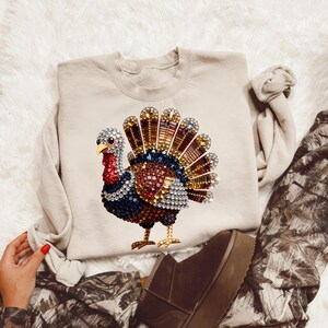 May include: A beige crewneck sweatshirt featuring a central graphic of a turkey composed of multicolored rhinestones. The turkey design displays a fanned tail with brown, gold, and blue crystals, a red wattle, and a body filled with dark blue and red gems.