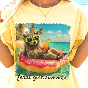 Puede incluir: Camiseta amarilla clara con un dise&ntilde;o de un mapache con gafas de sol en forma de coraz&oacute;n flotando en un flotador rosa en el mar, sosteniendo un c&oacute;ctel. El texto dice "feral girl summer".