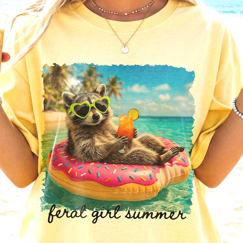 Puede incluir: Camiseta amarilla clara con un dise&ntilde;o de un mapache con gafas de sol en forma de coraz&oacute;n flotando en un flotador rosa en el mar, sosteniendo un c&oacute;ctel. El texto dice "feral girl summer".