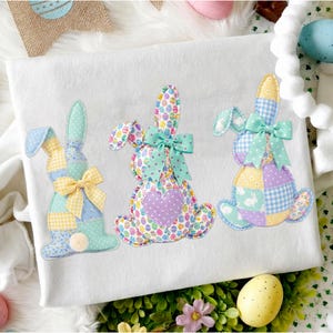 May include: White fabric with three appliqued Easter bunny designs. Each bunny is made of patchwork fabric in pastel colors, with bows and a pom-pom tail. The bunnies are decorated with egg and floral patterns.